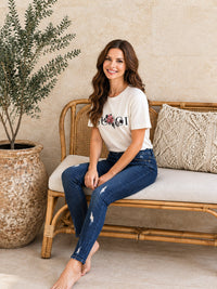 A woman sits on a woven rattan bench wearing a Villa Vibes white graphic t-shirt and dark blue jeans, set against a warm beige wall with a large potted plant.