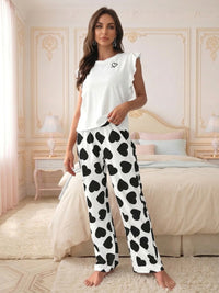 A woman modeling Villa Vibes Passion Princess two-piece pajama set in a bedroom. The set includes a white sleeveless top with a small black heart on the chest and matching white wide-leg pants covered in a black heart pattern.