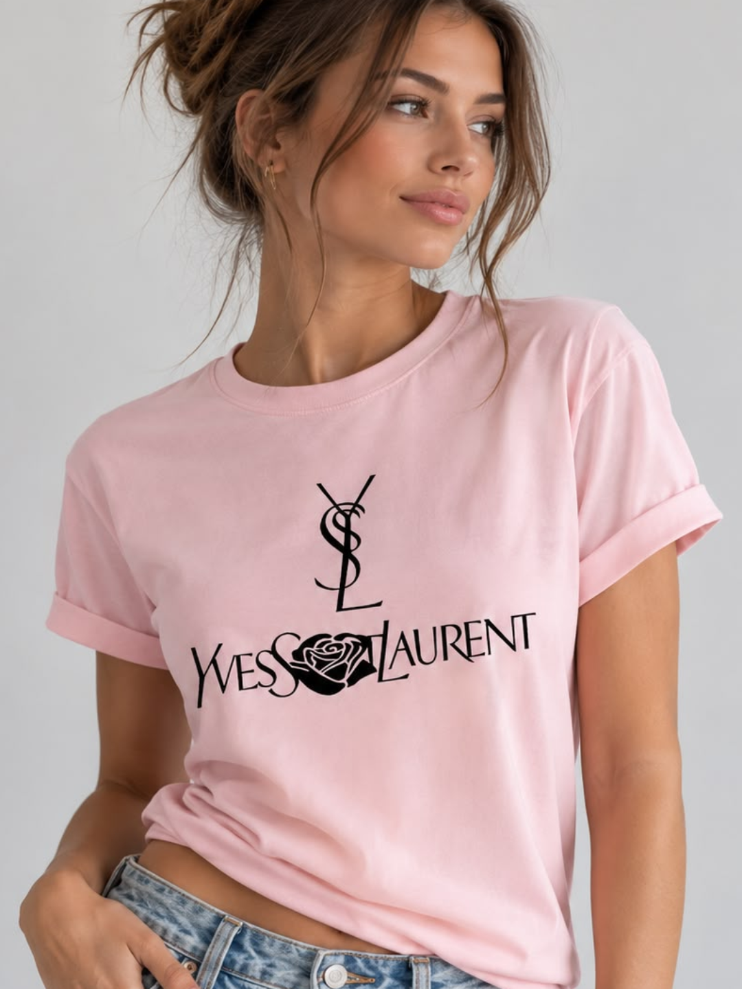 A woman wearing a Villa Vibes light pink short-sleeve t-shirt with a black printed Yves Saint Laurent logo and text on the front, paired with blue jeans, against a plain light gray background.