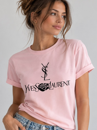 A woman wearing a Villa Vibes light pink short-sleeve t-shirt with a black printed Yves Saint Laurent logo and text on the front, paired with blue jeans, against a plain light gray background.