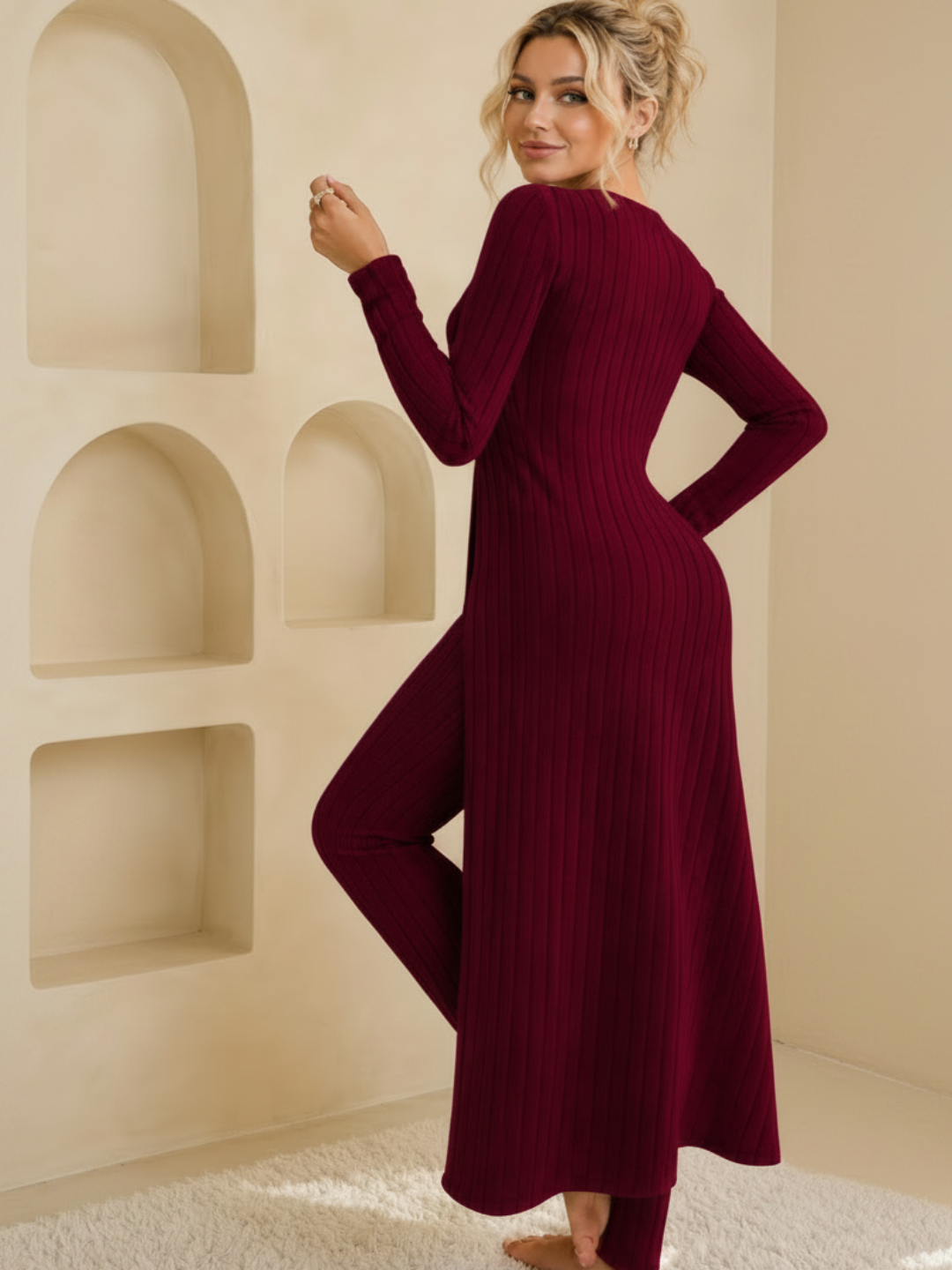 A woman wearing a Villa Vibes Shivani long-sleeve ribbed knit maxi dress in deep burgundy, standing in a warm beige room with recessed wall shelves.