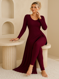 A woman wearing a Villa Vibes Shivani long-sleeve, ribbed knit maxi dress in deep burgundy, sitting on a modern beige stone pedestal in a minimalist room.