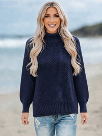 A woman with long blonde hair stands on a beach wearing a Villa Vibes Skye dark navy blue cable-knit mock neck sweater and light blue jeans.