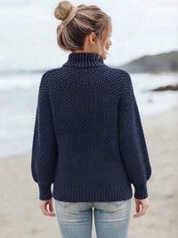 A woman with blonde hair in a bun stands on a beach, viewed from behind, wearing a Villa Vibes Skye dark navy blue textured knit turtleneck sweater and light blue jeans.