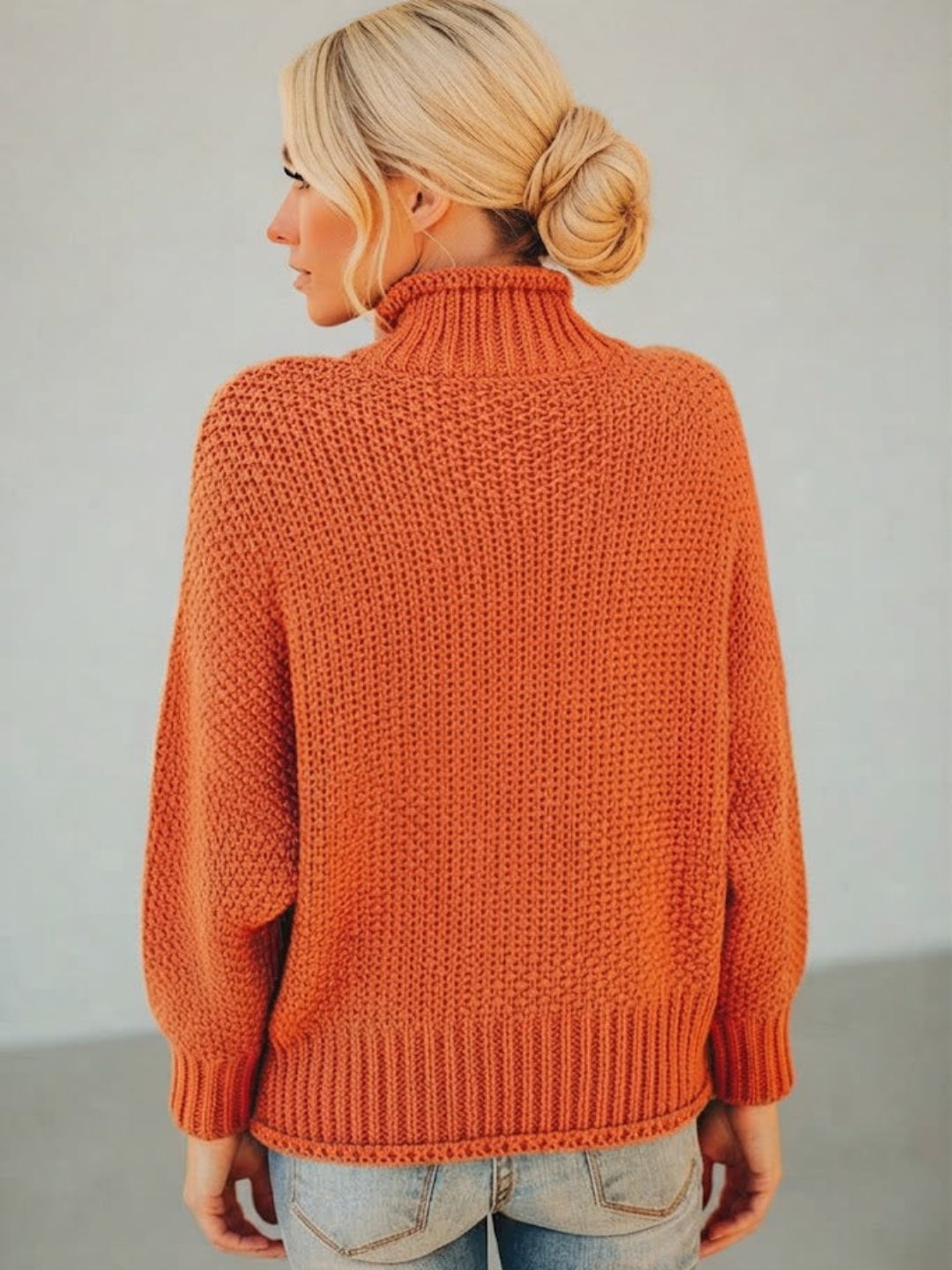 A woman with blonde hair in a bun is shown from the back wearing a Villa Vibes Skye chunky knit turtleneck sweater in a burnt orange color, paired with light wash jeans.