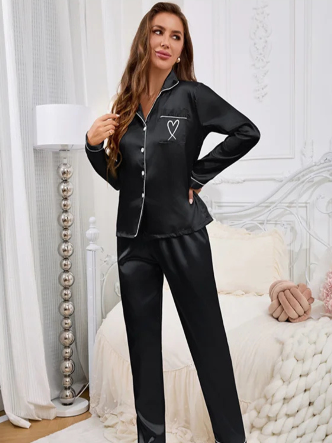 A woman wearing a Villa VIbes Sweet Dreams black silky two-piece pajama set with a heart design on the chest and white piping trim.