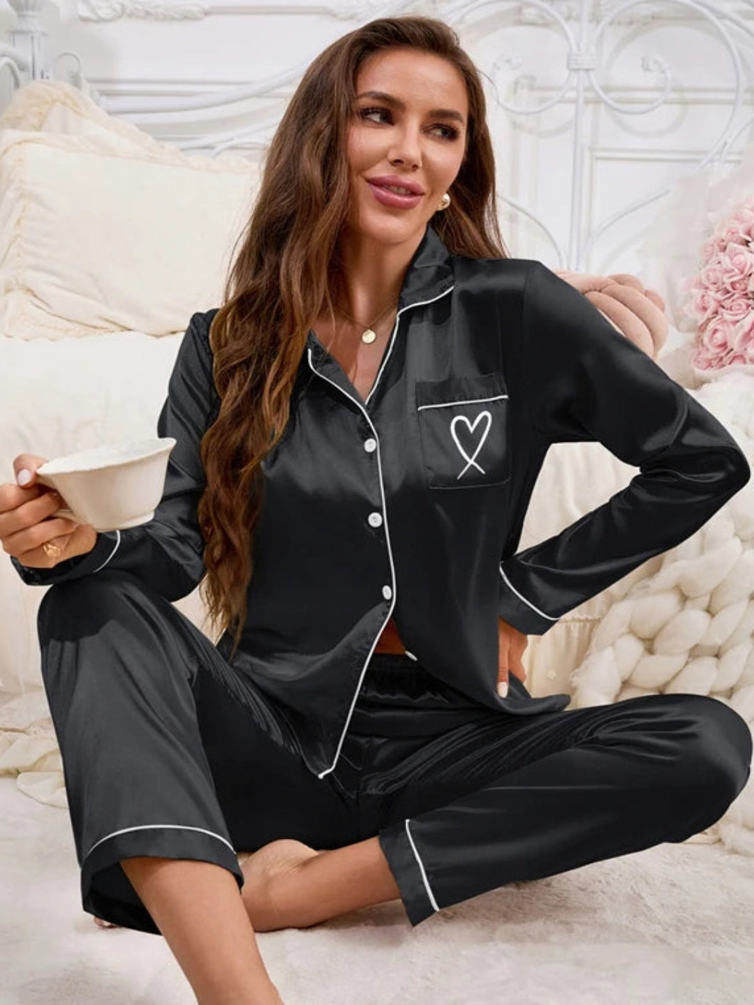 A woman wearing Villa Vibes Sweet Dreams black silky two-piece pajama set with a heart design on the chest and white piping trim.