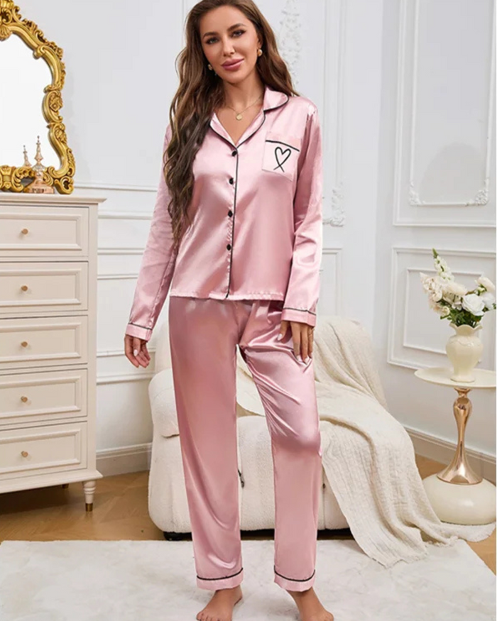 A woman wearing Villa Vibes Sweet Dreams light pink silky 2-piece pajama set with a turn-down collar, long sleeves, and pants. The pajama set features a black heart design pocket and elegant black piping trim.