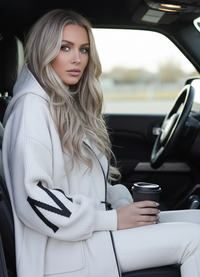 Woman sitting inside a car holding a coffee cup, wearing Kelly white coat with black accents.