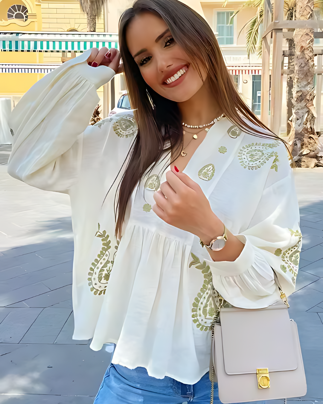 Woman wearing a Villa Vibes white blouse with green embroidery, holding a beige handbag on a street.