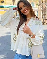 Woman wearing a Villa Vibes white blouse with green embroidery, holding a beige handbag on a street.