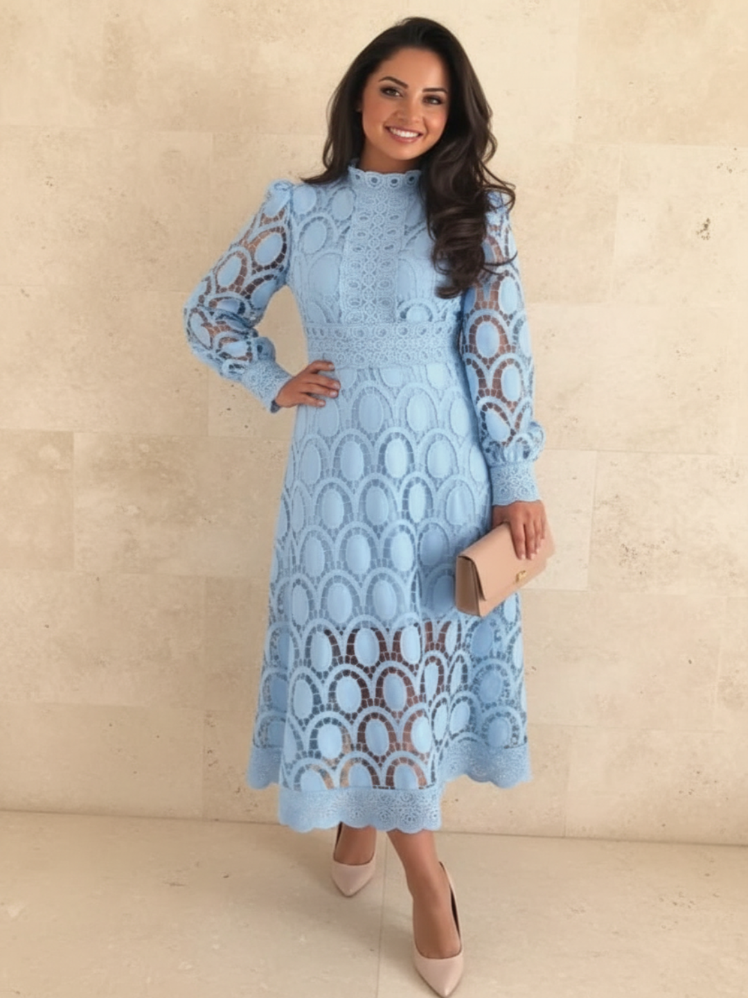 A woman models a Villa Vibes Jasmine light blue long-sleeve midi dress with an intricate lace and crochet pattern. She is holding a beige clutch and wearing beige heels, standing against a light beige stone wall.