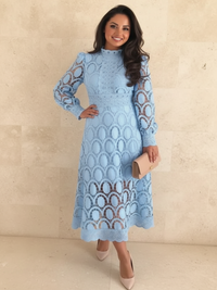 A woman models a Villa Vibes Jasmine light blue long-sleeve midi dress with an intricate lace and crochet pattern. She is holding a beige clutch and wearing beige heels, standing against a light beige stone wall.