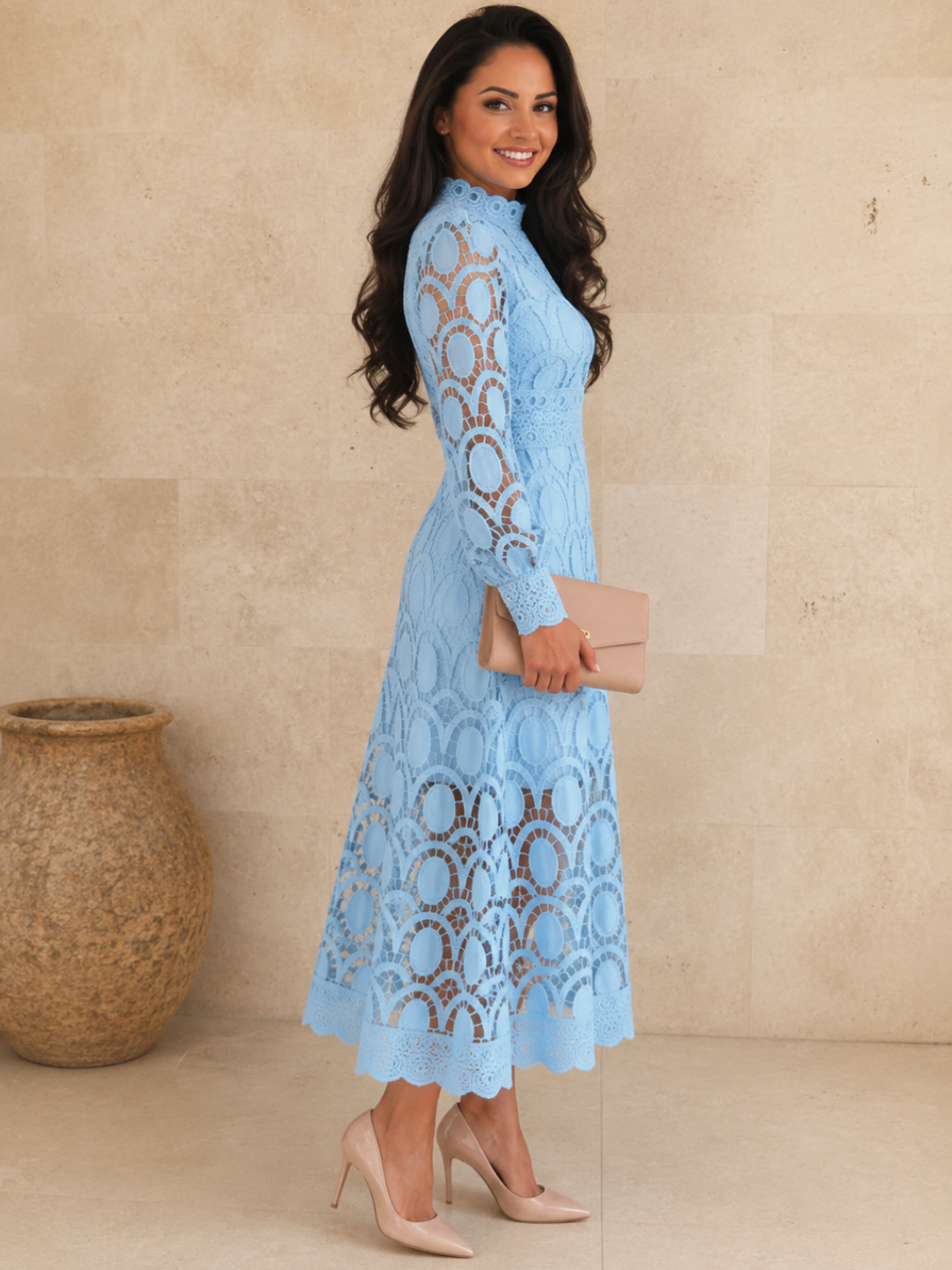 A woman modeling a Villa Vibes Jasmine light blue lace long-sleeve midi dress, holding a beige clutch and wearing beige high heels, standing against a beige stone wall.