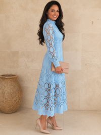 A woman modeling a Villa Vibes Jasmine light blue lace long-sleeve midi dress, holding a beige clutch and wearing beige high heels, standing against a beige stone wall.