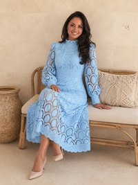A woman wearing aVilla Vibes Jasmine light blue long-sleeve lace midi dress sits on a woven rattan bench with a neutral textured wall and decorative ceramic pot in the background.